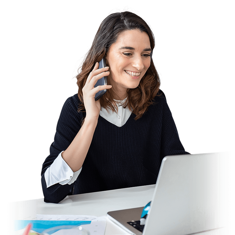 Eine mit dem Smartphone telefonierende Frau sitzt an einem Schreibtisch vor einem silbernen Laptop