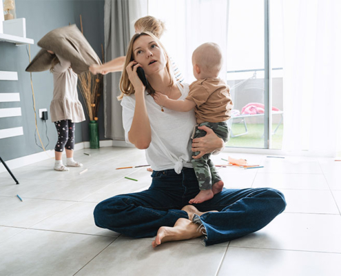 Gestresste Frau sitzt auf dem Boden, in einer Hand ein Smartphone am Ohr in der anderen ein Baby, während im Hintergrund zwei Kinder spielen