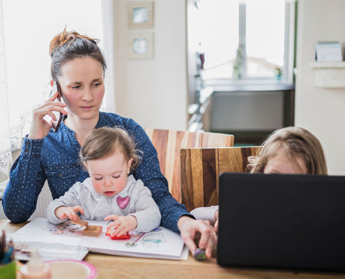 Mutter sitzt mit zwei Kindern am Küchentisch und telefoniert während das jüngere bei ihr auf dem Schoss sitzt und spielt und das andere vor einem Laptop sitzt – vitaL
