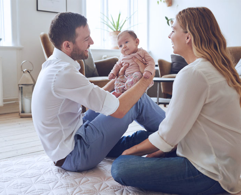 Eine junge Familie sitzt auf dem Boden im Wohnzimmer und der Vater hält das Kind auf seinen Knien – vitaL