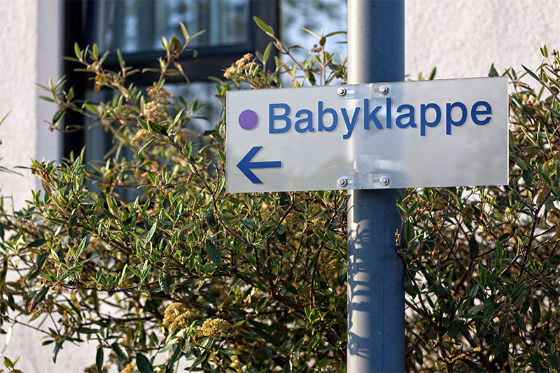 Das Schild einer Babyklappe befestigt an einem Laternenmast vor einer Hecke