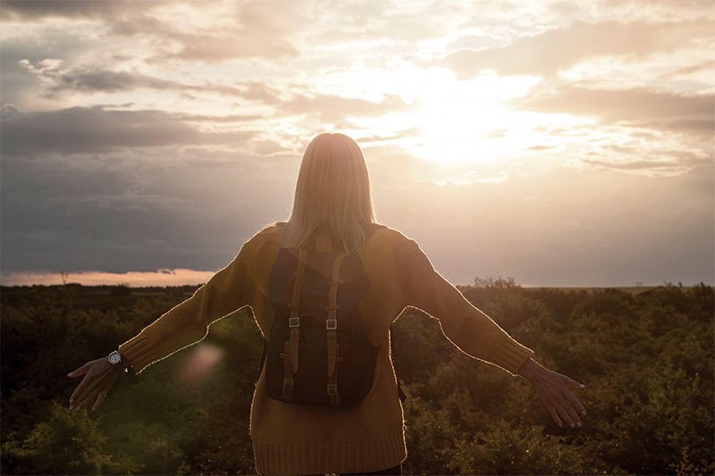 Eine junge Frau in gelbem Pulli & mit Rucksack steht im Sonnenuntergang auf einer Anhöhe vor einem Wald richtung Sonne gewandt und hat die Arme ausgebreitet